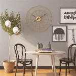 Relógio de parede com estrutura de metal dourado, design geométrico, numa sala com quadros decorativos e uma planta em vaso de barro.