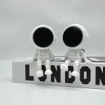 Duas figuras de astronautas em estilo minimalista, com cabeças grandes e corpos pequenos, sentados numa placa com a palavra 'LONDON' em letras pretas.