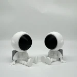 Dois robôs de aparência futurista, com cabeças grandes e corpos pequenos, sentados no chão com as pernas esticadas, em fundo branco.