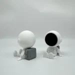 Dupla de figuras de astronauta com cabeças grandes, uma com rosto preto e outra com cabeça branca, sentadas em fundo branco.