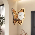 Relógio de parede em forma de borboleta com detalhes decorativos e iluminação de fundo, colocado numa parede clara ao lado de uma janela com persianas.