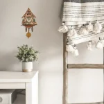Relógio de parede estilo chalé com detalhes em madeira, ao lado de uma planta em vaso branco e uma cortina de tecido branco com pompons, em ambiente de casa.