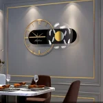 Relógio de parede com mostrador de 12 horas, detalhes dourados e elementos decorativos em preto e branco, instalado numa parede cinzenta com moldura dourada.