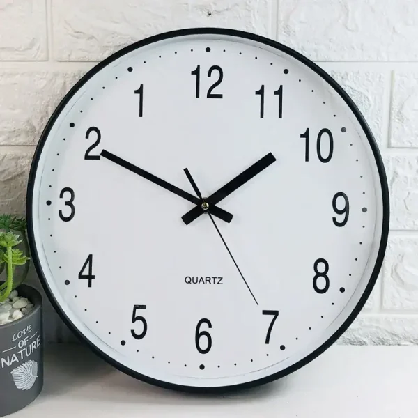 Relógio de parede com mostrador branco, números pretos e ponteiros pretos, exibindo a hora 10:10, com fundo de parede de tijolos brancos e uma planta ao lado.