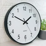 Relógio de parede com mostrador branco, números pretos e ponteiros pretos, exibindo a hora 10:10, com uma planta ao lado.