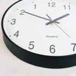Relógio de parede com mostrador branco, números pretos e ponteiros cinzentos, exibindo a hora 2:10:36.