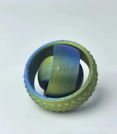 Bola de borracha verde com textura rugosa, contendo uma esfera de metal com acabamento colorido dentro, vista de cima.
