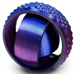 Anel de metal com acabamento em azul e roxo, com textura rugosa na borda e superfície lisa no interior, visto de ângulo lateral.