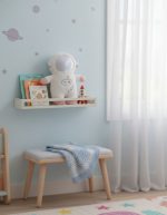 Almofada de astronauta de peluche em prateleira com livros infantis e foguete decorativo, numa parede com tema espacial, em quarto de criança bem iluminado.