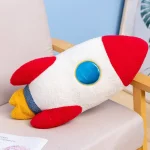Pelúcia de foguete com corpo branco, detalhes vermelhos, azuis e amarelos, colocada numa cadeira de sala de estar.