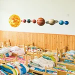Mural com representação do sistema solar, incluindo o Sol, planetas e anéis de Saturno, decorando uma sala com várias camas de madeira e roupas de cama coloridas.