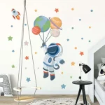 Adesivo de astronauta com planetas e foguete na parede de quarto infantil, com balanço de madeira e luminária de leitura ao lado.