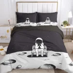 Conjunto de roupa de cama com estampa de astronauta sentado numa lua, com estrelas e crateras, em cores preto e branco, numa cama de quarto infantil.