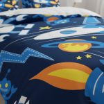 Capa de edredão com desenhos de planetas, estrelas, foguetes e astronautas em cores vibrantes sobre fundo azul escuro.