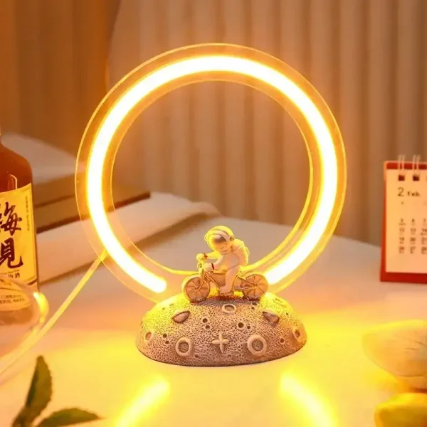 Lâmpada de mesa com anel de luz LED amarelo, base decorativa com figura de criança a andar de bicicleta, ambiente acolhedor com calendário e garrafa ao fundo.