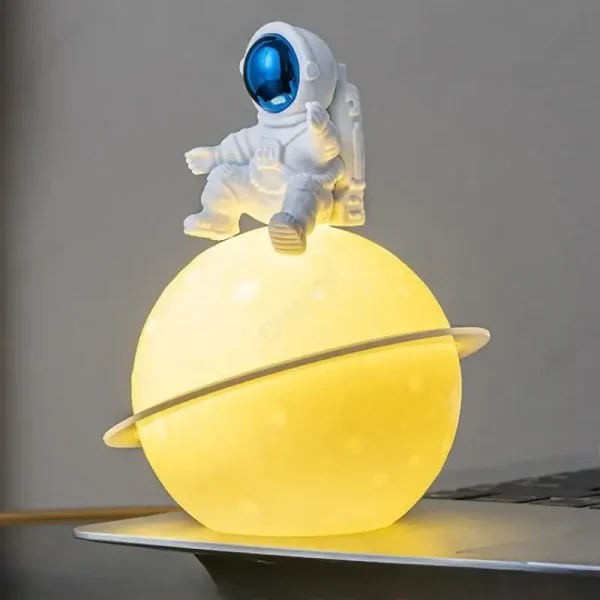 Luminária de planeta amarelo com anel, com figura de astronauta sentado na parte superior, em ambiente de decoração científica.