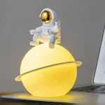 Luminária de planeta amarelo com astronauta em traje espacial branco com visor dourado, sentado na parte superior do planeta, decorativa para espaço ou ciência.