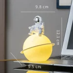 Lâmpada decorativa em forma de planeta com astronauta em cima, iluminação amarela, dimensões indicadas, ao lado de um computador portátil.