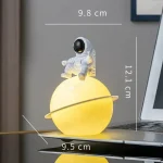 Luminária decorativa de planeta com astronauta sentado na parte superior, iluminação amarela suave, dimensões visíveis de 9.8 cm de altura e 9.5 cm de diâmetro.