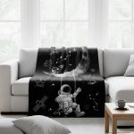 Manta preta com ilustrações de astronauta, planetas, estrelas e foguetes, com uma grande lua ao centro, colocada sobre um sofá branco em sala bem iluminada.