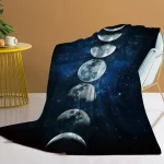 Manta de tecido com impressão das fases da Lua, colocada sobre uma cadeira de descanso, com fundo de parede bege e mesa amarela ao lado.