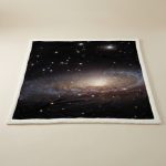 Manta com imagem da galáxia Andrómeda, detalhes de estrelas e nebulosas, vista de perto, com borda branca.