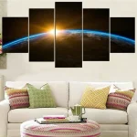 Painel decorativo de cinco painéis com imagem da Terra vista do espaço e o Sol nascente, colocado acima de um sofá branco com almofadas coloridas.