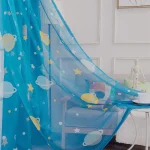 Cortina de cama azul com desenhos de planetas, estrelas e satélites, decorando um quarto infantil com móveis brancos e parede decorada com molduras.