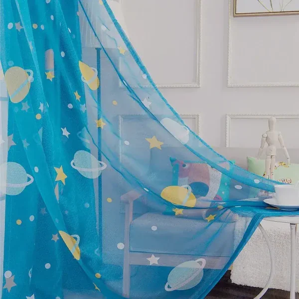 Cortina de cama azul com desenhos de planetas, estrelas e satélites, decorando um quarto infantil com móveis brancos e parede decorada com molduras.
