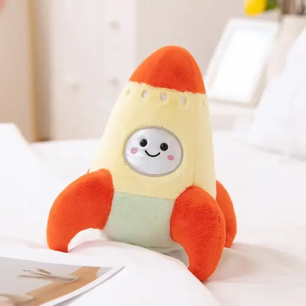 Boneco de peluche com corpo de foguete, rosto sorridente, detalhes em cores laranja, amarelo e verde, colocado numa cama com fundo de quarto.