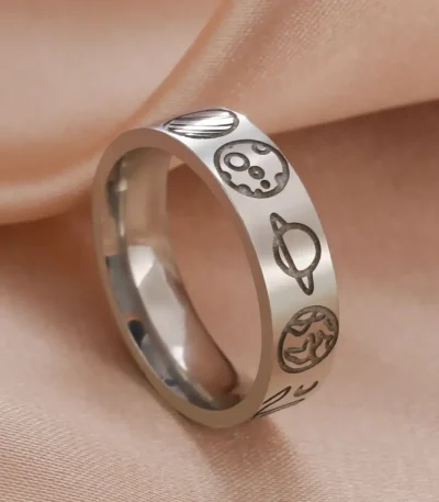 Anel de metal prateado com símbolos de planetas e estrelas gravados, colocado sobre tecido de cor pêssego.