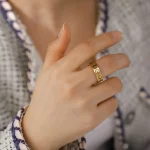 Close-up de uma mão feminina com anel de ouro com símbolos gravados, usando roupa de tecido com padrão de linhas e cores neutras.