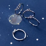 Três anéis de prata com detalhes brilhantes, um deles com uma pedra central, colocados sobre fundo azul com estrelas e uma lua dourada.