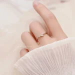 Anel de diamantes com detalhes brilhantes na mão de uma pessoa, vestindo roupa branca de manga comprida, fundo neutro.