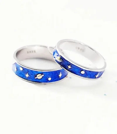 Dois anéis de prata com decoração de espaço, incluindo planetas, estrelas e uma nave espacial, com fundo branco.