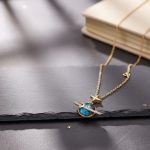 Colar fantasia com pingente de planeta e estrela, inspirado no espaço, em ouro e azul, colocado sobre uma superfície escura com um livro ao fundo.