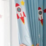 Cortina azul com desenhos de foguete vermelho, estrelas amarelas e detalhes em cinza, decorando uma janela de quarto infantil.