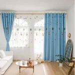 Cortinas azuis com detalhes de foguetes, estrelas e planetas, em uma sala bem iluminada com janelas grandes e decoração infantil.
