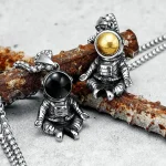 Pingentes de astronautas em metal com bolas de cor dourada e preta, suspensos em uma corrente, inspirados no espaço e na exploração espacial.