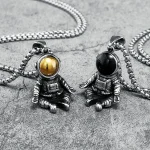 Pingentes de astronautas em metal com esferas dourada e preta, suspensos em correntes, concebidos como joias originais e modernas inspiradas no espaço.