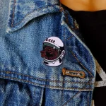 Pin original em forma de cão usando um capacete de astronauta com inscrição "CCCP", fixado numa jaqueta de ganga azul, estilo casual.