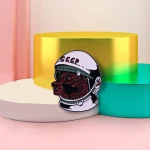Pingente em forma de cabeça de cão usando um capacete espacial com inscrição "CCCP", colocado sobre suportes coloridos em tons pastel.