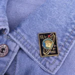 Prendedor de gravata em metal com motivo espacial e data de 1961, fixado numa camisa de ganga azul clara, adicionando um toque de estilo vintage e científico ao visual.