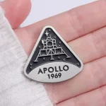 Badge em forma de triângulo representando o módulo lunar Apollo 1969, com detalhes em relevo e inscrição "APOLLO 1969", ideal para colecionadores e entusiastas do espaço.