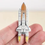 Pin em forma de nave espacial com detalhes em metal, ilustrando um foguete pronto para decolar, perfeito para os amantes de astronomia e ciência.
