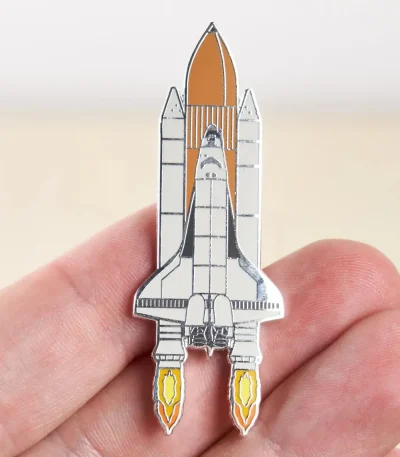 Pin em forma de nave espacial com detalhes em metal, ilustrando um foguete pronto para decolar, perfeito para os amantes de astronomia e ciência.