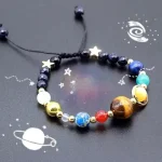 Pulseira com contas representando planetas do sistema solar, estrelas e elementos metálicos, sobre fundo com desenhos de galáxias e planetas.