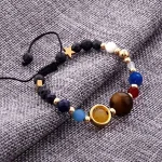 Pulseira com contas de diferentes cores e tamanhos, incluindo azul, branco, dourado e uma grande bola de madeira, sobre tecido cinza.