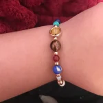 Pulseira com contas de diferentes cores e materiais, incluindo azul, marrom, vermelho e branco, com fecho dourado, usada no pulso de uma pessoa.