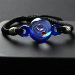 Pulseira de cordão negro com esfera de vidro azul e detalhes metálicos dourados.